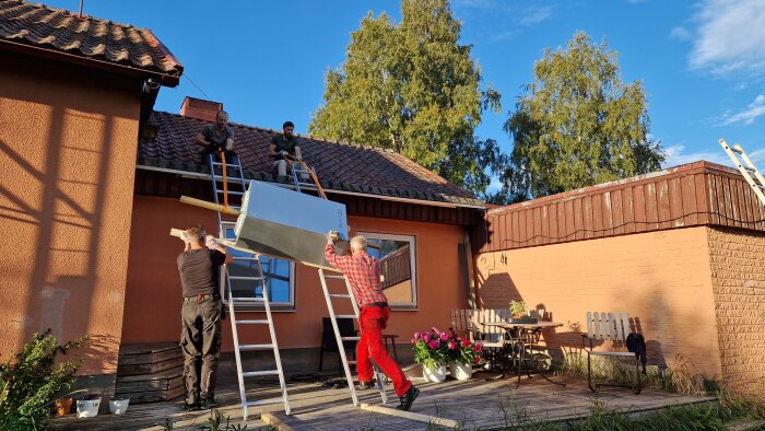 Personer flyttar ett ventilationsaggregat uppför en stege till ett tegeltak på ett orange hus. Två personer hjälper till från taket.