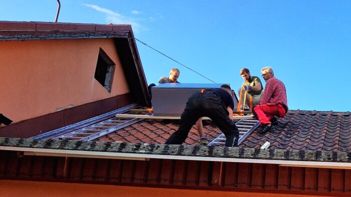 Fyra personer installerar ett ventilationsaggregat på ett tegeltak, använder stegar och remmar för att hantera utrustningen försiktigt.