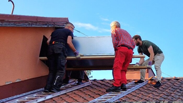 Tre personer lyfter ett ventilationsaggregat genom ett vindsfönster på ett tegeltak med hjälp av stegar för att stötta processen.