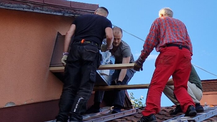 Fyra personer lyfter ett ventilationsaggregat via ett vindsfönster på ett tegeltak för installation.