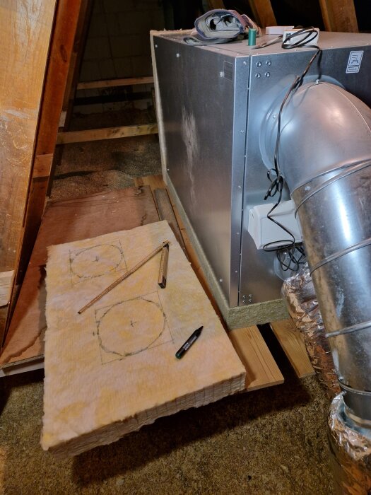 Ventilationsaggregat med ansluten metallisk ventilationskanal på vinden, omgiven av isoleringsskivor och verktyg, inklusive en tumstock och en penna.