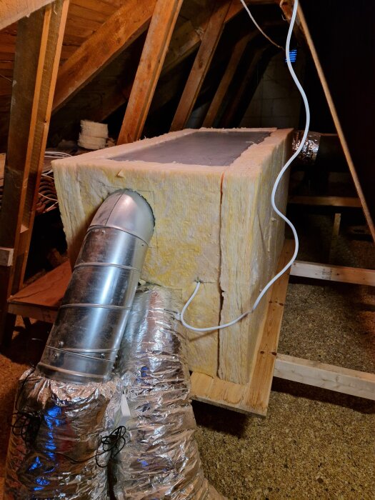 Isolerad ventilationskanal på vind, med silverfärgade flexrör och elledning, monterad i ett trästativ.