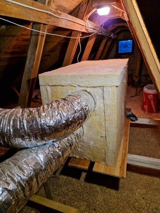 Isolerad ventilationslåda med två metalliska rör i en vindsvåning, upplyst av en lampa, med trätak och fönster i bakgrunden.