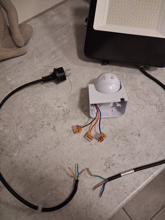 Elektrisk installation pågående, kablar anslutna till rörelsesensor och kopplingsblock; strömsladd och lampa synliga på arbetsbänk.