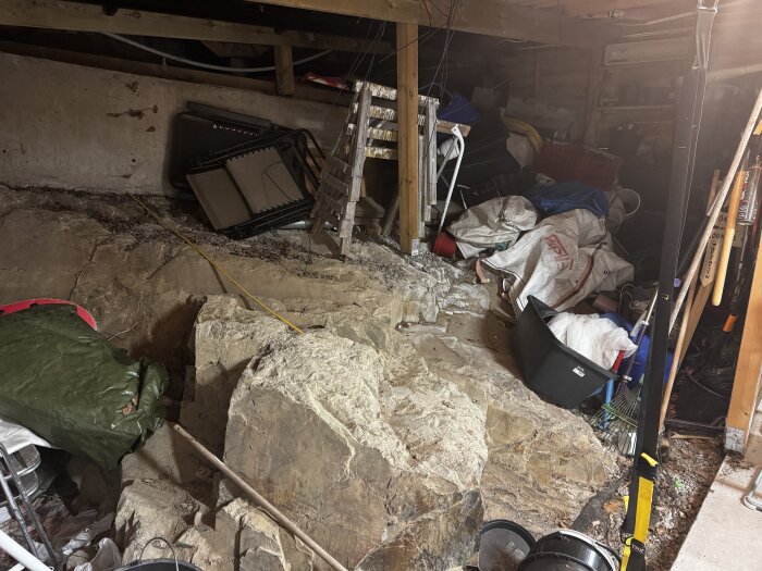En källare med oorganiserad förvaring, stenar på marken, byggmaterial och verktyg staplade mot väggen.