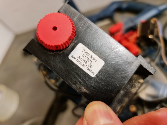 Svart varvtalsregulator med en röd ratt, märkt "230V 50Hz", hållen i handen. Synlig text och bakgrund med oskarpa verktyg.