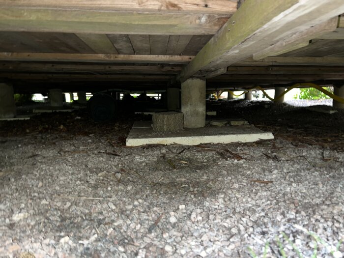 Grundbjälkar och torrt virke under ett hus, med grusad mark för att förhindra fukt.