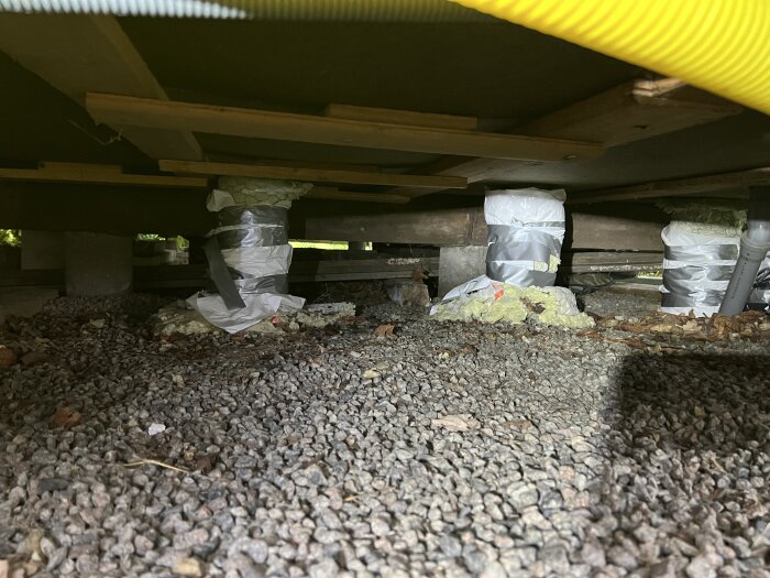 Under huset med grusad mark, syns bjälkar av icke-tryckt virke, stöd i plast och tejp, samt torrt trä.