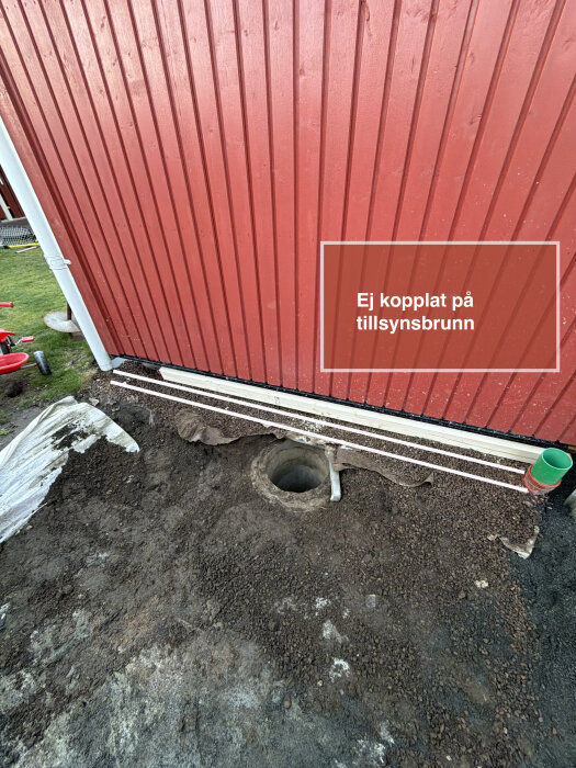 Perforerat dräneringsrör vid husgrund, ej kopplat till tillsynsbrunn. Grus och jord omkring. Rött hus och vit stupränna syns i bakgrunden.