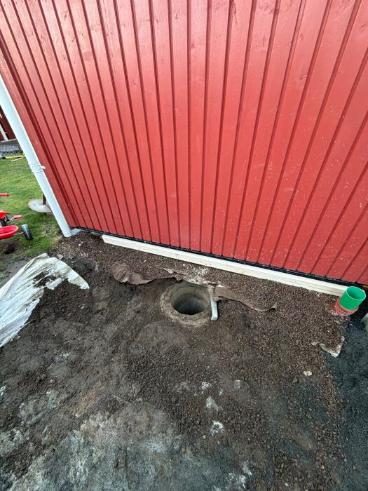 Perforerat rör vid husgrund, omgivande jord och grusstensbädd, bredvid röd trävägg; del av projekt för gjutning av genomfart.