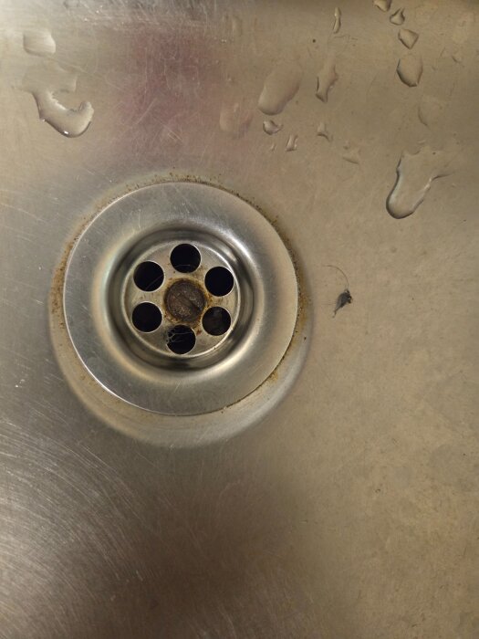 Närbild av ett diskbänksavlopp i metall utan propp, med vattenstänk och åtta runda hål runt centrum.