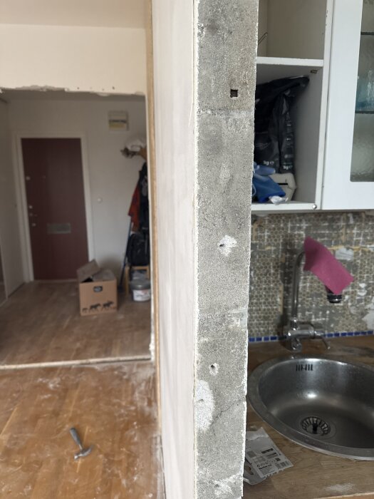 Lättbetongvägg under renovering med borrade hål, del av kök med diskbänk och mosaik i bakgrunden, samt en kartong och verktyg på golvet.
