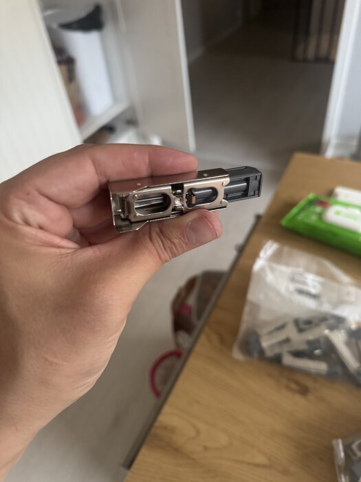 Närbild på en person som håller en Blum-dämpare för köksluckor, mot en bakgrund med ett bord och påsar.