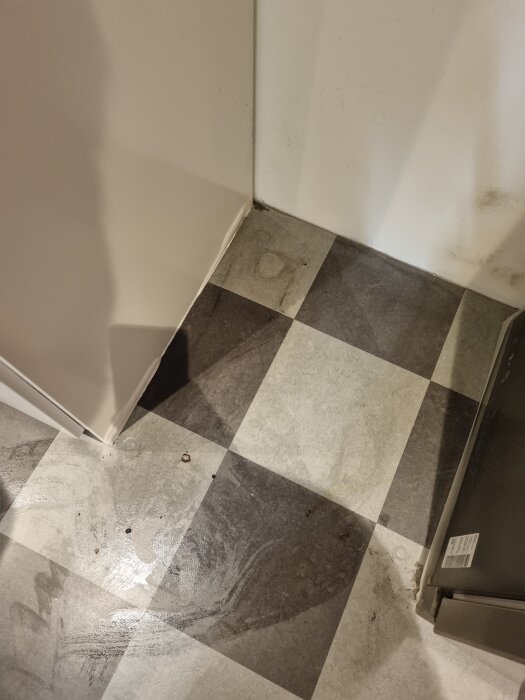 Svartvitt rutigt golv i ett hörn med smutsiga märken och fläckar. Skåpväggen i vitt syns på vänster sida.