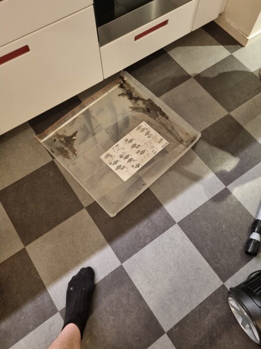 Ugnslucka med glasskiva på ett rutigt köksgolv, instruktioner ligger ovanpå och en person med svart strumpa syns bredvid.