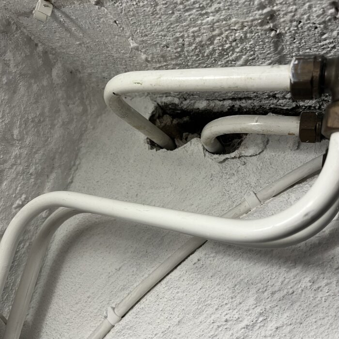 Vita rör i en vägg med oregelbunden yta och hål, användaren undrar om materialet kan vara asbest.