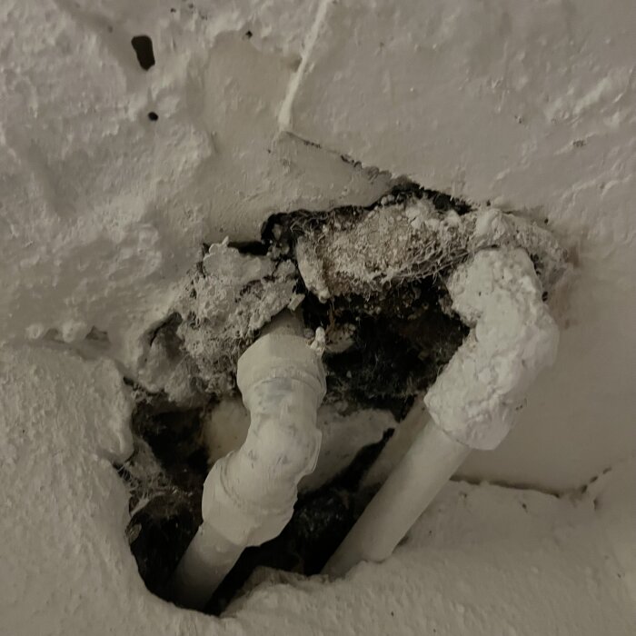 En närbild av gamla metallrör och trasig isolering i en vit vägg, potentiellt misstänkt för asbestinnehåll.