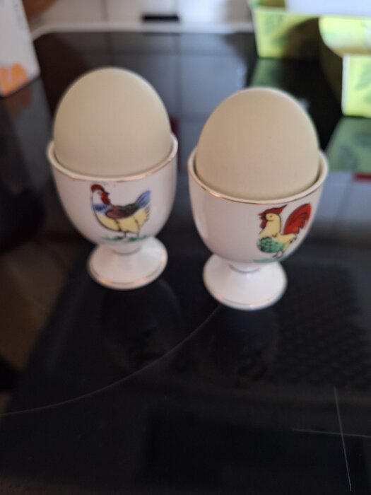 Två ägg i äggkoppar med tuppmotiv, placerade på en mörk köksbänk.