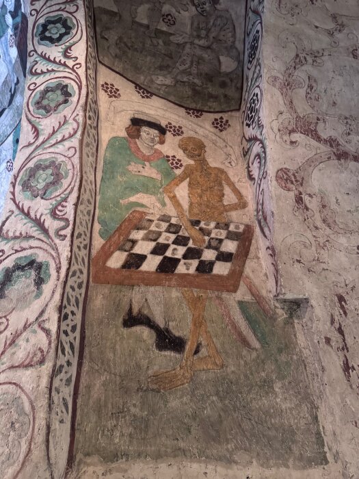 Målning av två figurer som spelar schack; en levande man och ett skelett. Bordet har svartvita rutor. Dekorativa blommönster omger motivet.