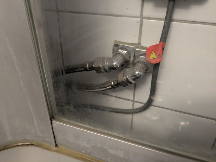 Rörkopplingar och slangar till duschblandare på en kaklad vägg, med en varningsetikett på en av slangarna.