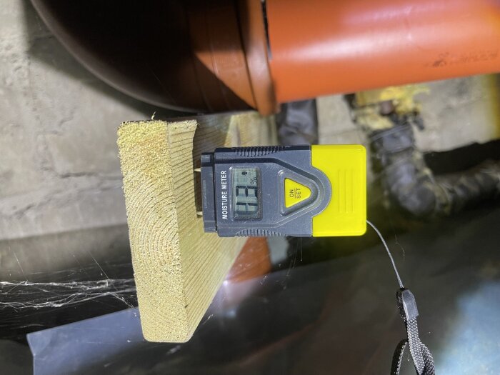 Fuktmätare placerad på en träregel i en krypgrund, visar 11,9% fuktighet med en orange ventilationsrör synligt i bakgrunden.