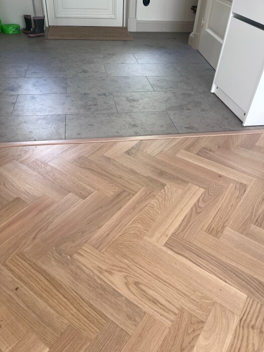 Parkettgolv i fiskbensmönster möter grått klinkergolv i en hall. Hallen har vita väggar och stövlar vid väggen.