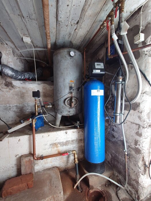 Gammal hydrofor och blå filtertank i ett källarutrymme med röranslutningar och ventil. Taket och väggarna är rustika och slitna.