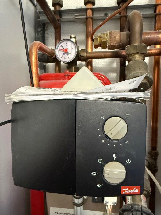 Styrpanel för värmesystem med två vred (+8 till -8 och sol-symbol), kopparrör, manometer och Danfoss-logotyp.