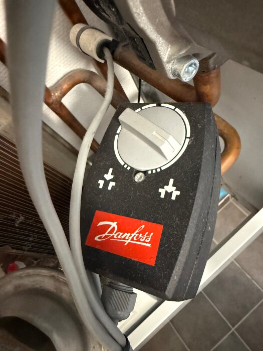 Danfoss värmeregulator med ratt och plus/minus-symboler, ansluten till rör och elkabel; del av värmesystem i hus.