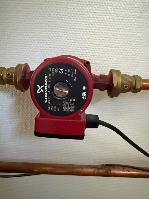 En röd Grundfos cirkulationspump, modell UPS 25-40, kopplad till kopparrör och placerad mot en vit vägg.