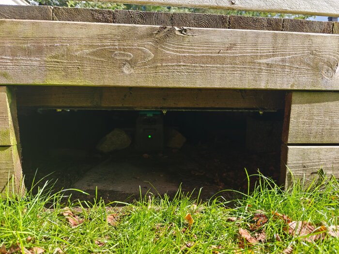 Laddstation under trädäck skyddad av akrylplastskiva, synlig genom öppning i träkonstruktionen. Gräs och löv i förgrunden.
