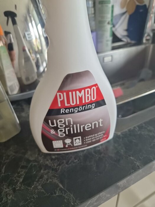 Flaska med Plumbo rengöring för ugn och grillrent står på en mörk bänkskiva i ett kök, omgiven av andra rengöringsmedel.