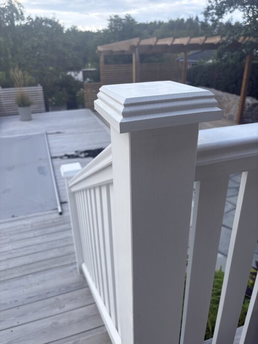 Vitmålat trappräcke och stolpe i närbild på en altan med trätrall och en pergola i bakgrunden.