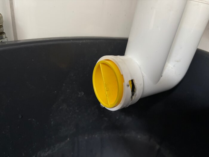 Vit rördel med gul plastpropp monterad ovanpå en svart behållare i ett installationsutrymme.