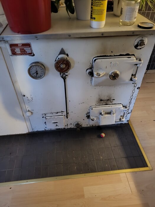 Gammal vedpanna med reglage och mätare, placerad på ett klinkergolv med en röd hink och flaskor ovanpå.