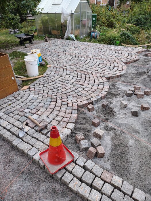 Stenläggning pågår i trädgård med halvfärdigt mönster av stenar; röd kon och verktyg syns, växthus i bakgrunden.
