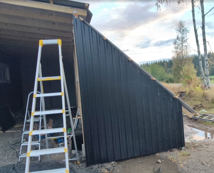 Svart plåtvägg monterad på byggprojekt, med en stege framför. Utspridda verktyg och material på marken. Skog och himmel i bakgrunden.