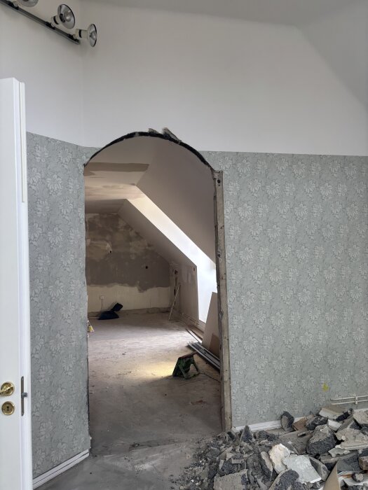 Öppning i vägg med halvfärdigt valv och mönstrad tapet, oavslutat arbete och bråte på golvet, belysning i taket.