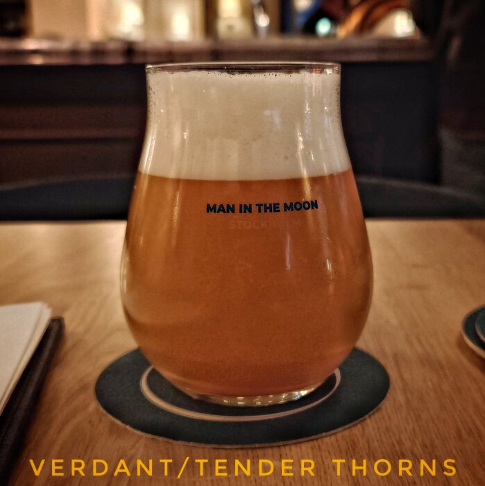 Ett glas öl från Man in the Moon med skumkrona, placerat på ett bord i en bar. Texter på glaset och bordet.
