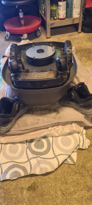 Robotgräsklippare Husqvarna Automower uppochnervänd för rengöring, placerad på en filt bredvid ett par svarta skor i ett garage.