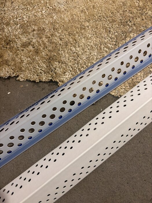 Två perforerade hörnlister av plast och metall, placerade mot en bakgrund av betong och spackel, för användning vid skarpa ytterhörn.