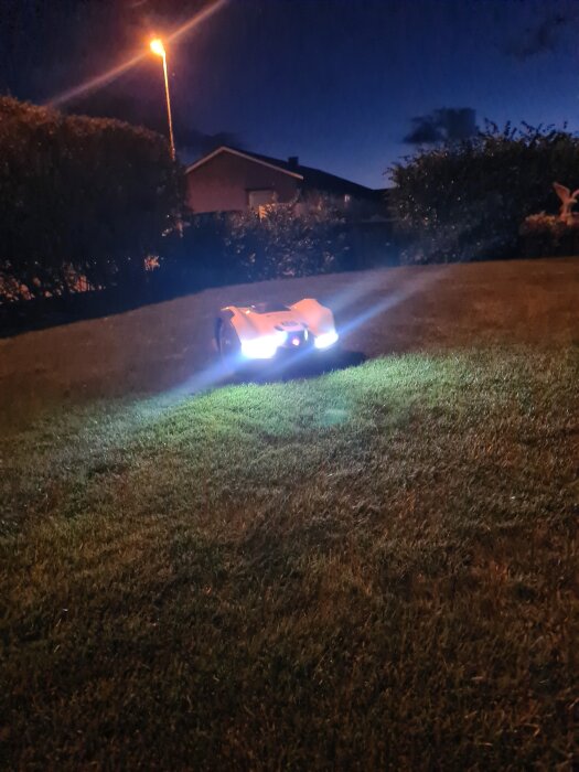 Robotgräsklippare Husqvarna Automower på natten med lysande lampor, klipper gräs framför en upplyst trädgård i mörker.
