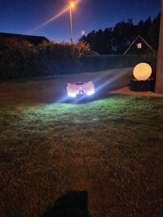 Robotgräsklippare Husqvarna Automower på en mörk gräsmatta med lysande strålkastare, mot en bakgrund av buskar och hus i skymning.