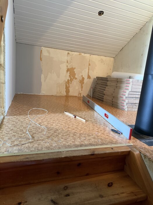 Liten hall på övervåning i timmerhus med ojämnt golv som visas tillsammans med vattenpass, golvpaket och avskalad tapet.