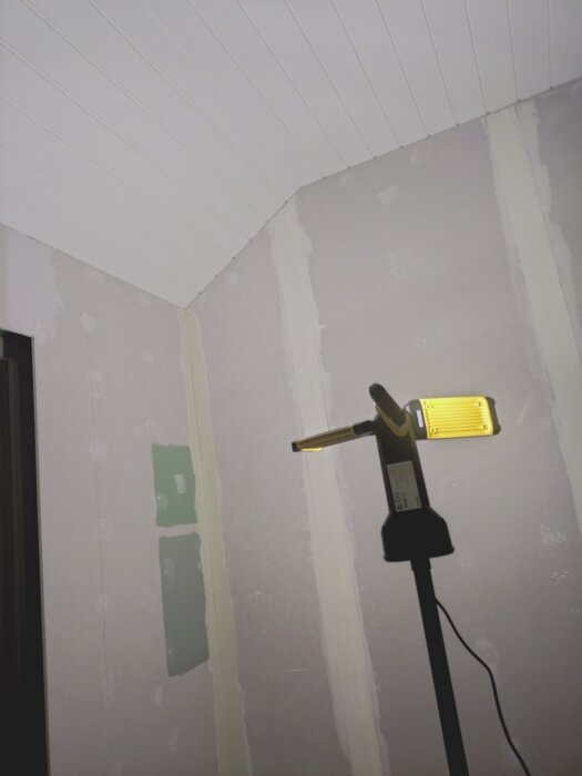 Arbeidslampa lyser opp et rom med uferdige vegger og hvitt takpanel.