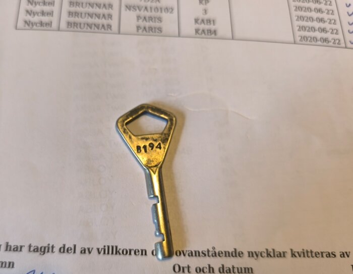 Metallnyckel märkt B194 liggande på ett papper med text och kolumner. Nyckeln har en sexkantig topp och är fotograferad ovanifrån.