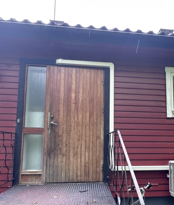 Ytterdörr med luftvärmepump och kanallist installerad ovanför, som sticker ut från fasaden på ett rött trähus.