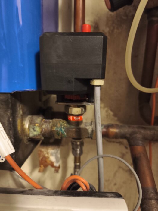 En detaljbild av ett rörsystem med ventiler och kopplingar, delvis målade i blått, och en svart låda med en röd knapp ovanpå.