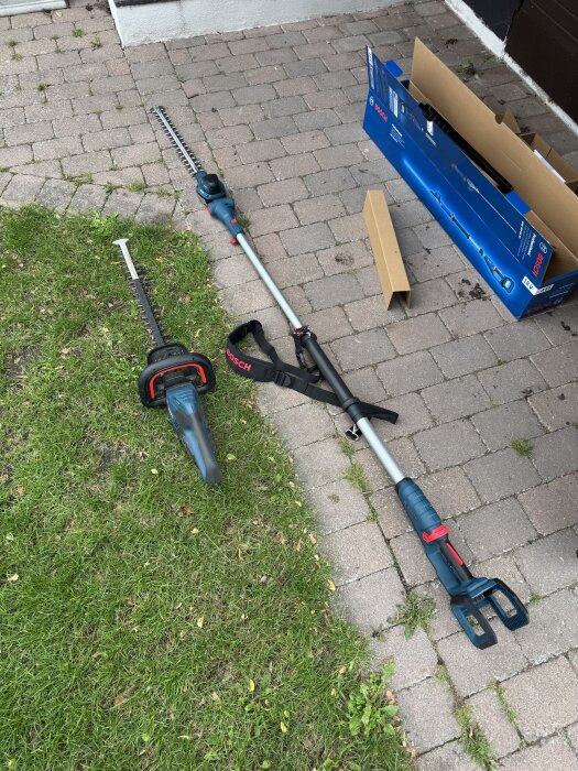 Häcksax med långt skaft ligger på stengolv bredvid förpackning och kartong, delvis på en gräsmatta.