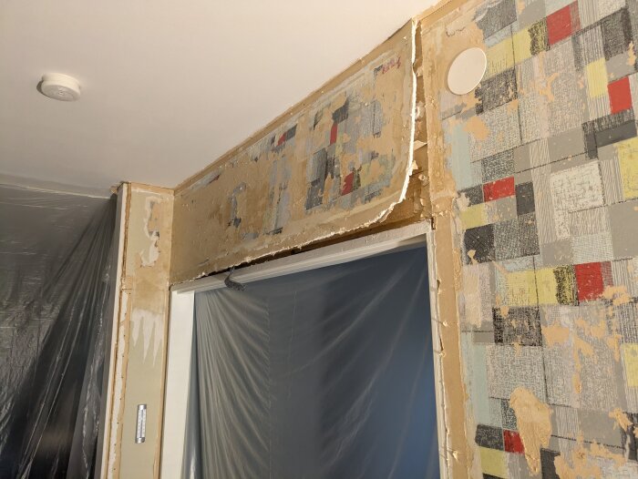 Rivet gips på vägg med färgglada rester, dammigt utrymme. Plastskynken täcker dörröppningar, pågående renovering synlig.